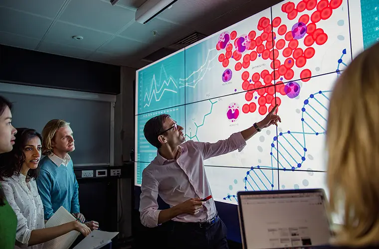Ein Lektor deutet auf einen Bildschirm mit roten Punkten und der Skizze einer DNA während 4 Personen ihm zuhören (Foto)