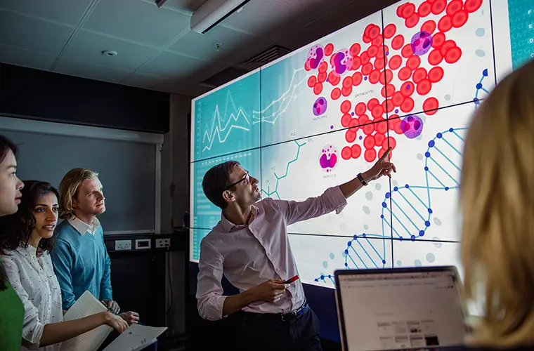 A lector is pointing at a screen with red dots and the sketch of a DNA while four people are watching him (Photo)