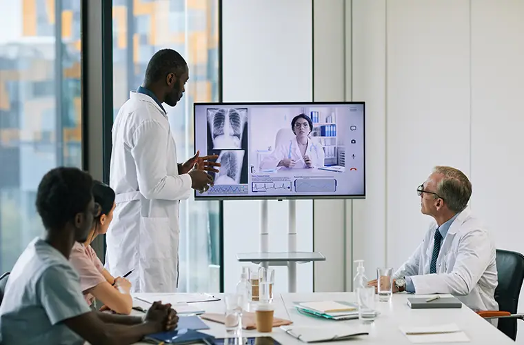A doctor is holding a presentation in front of three people sitting at a table and is pointing at a TV screen. (Photo)