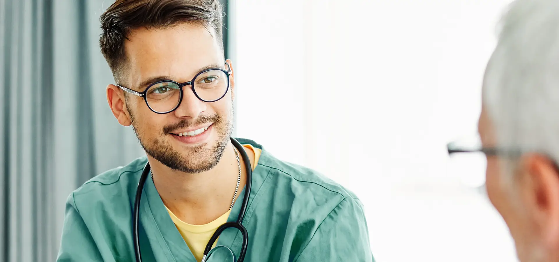 A doctor in green scrubs is talking to a patient; view over the shoulder of the patient (Photo)